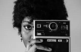 Schwarzweiß-Foto einer Schwarzen Frau, die eine alte Kamera vor ihr linkes Auge hält
