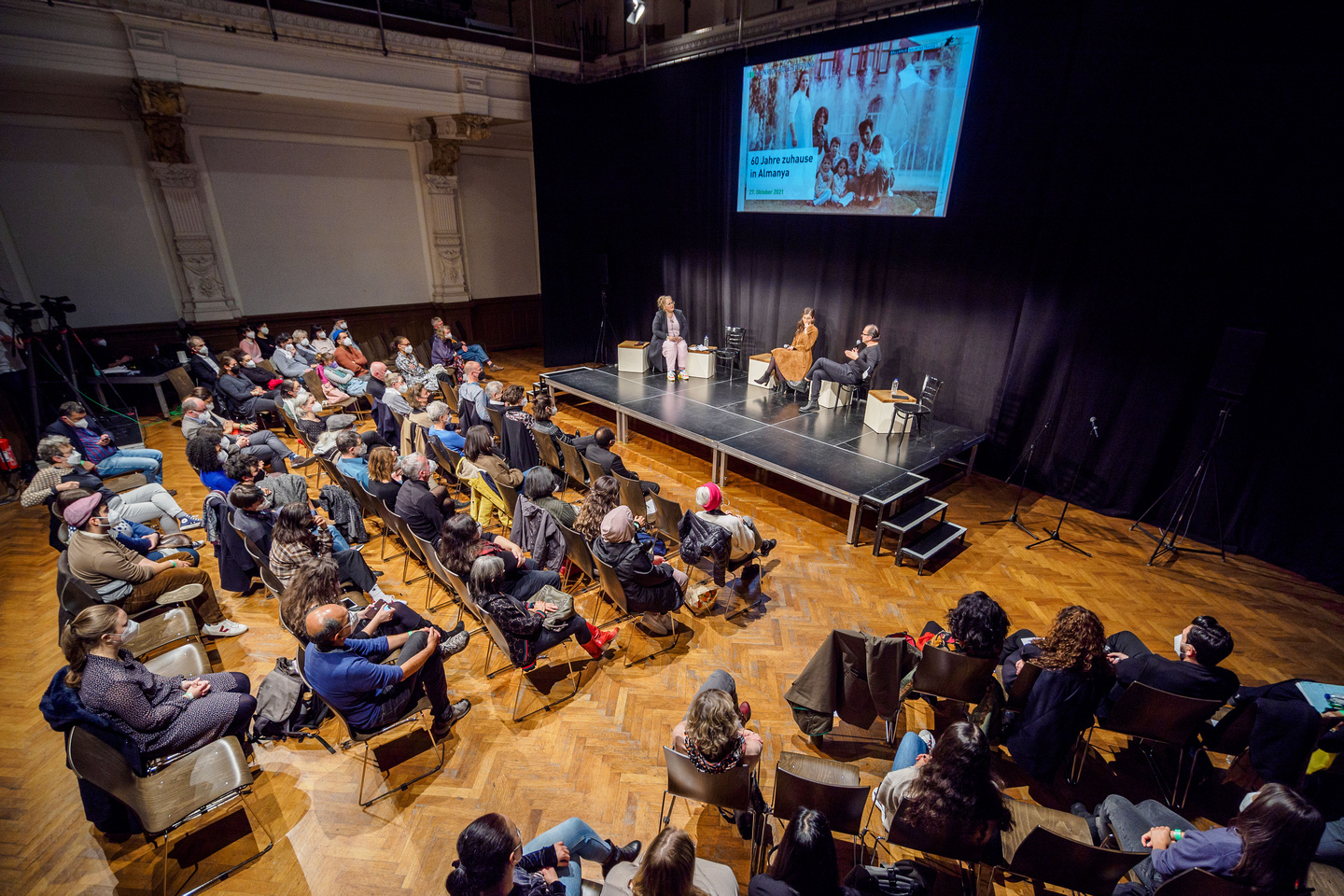 60 Jahre zuhause in Almanya Veranstaltung 60 Jahre zuhause in Almanya, zu sehen ist der Theatersaal im Ballhaus Naunynstraße mit Publikum sowie das Podium mit Idil Baydar, Tuba Bozkurt und Imran Ayata.