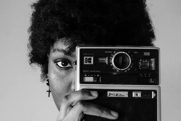 Schwarzweiß-Foto einer Schwarzen Frau, die in die Kamera schaut und eine alte Kamera vor ihr linkes Auge hält