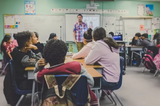 Kinder sitzen in einer Schulklasse, im Hintergrund steht ein Lehrer an der Tafel