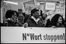 Bei einer Demo halten Schwarze Menschen einen Banner mit der Aufschrift "N*Wort stoppen!", im Hintergrund sind weitere Demoschilder zu sehen, mit der Aufschrift "#Black" oder "Wir erheben Einspruch".