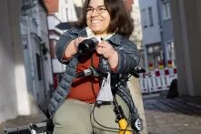 Foto von Andrea Schöne, die lächelnd auf einem Rad sitzt und eine Fotokamera in der Hand hält