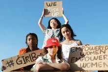 Vier junge Frauen schauen selbstbewusst in die Kamera und halten Schilder mit folgender Aufschrift hoch: "We rise up", "Girlboss", "Empowered women empower women"