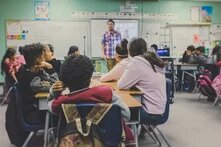 Kinder sitzen in einer Schulklasse, im Hintergrund steht ein Lehrer an der Tafel