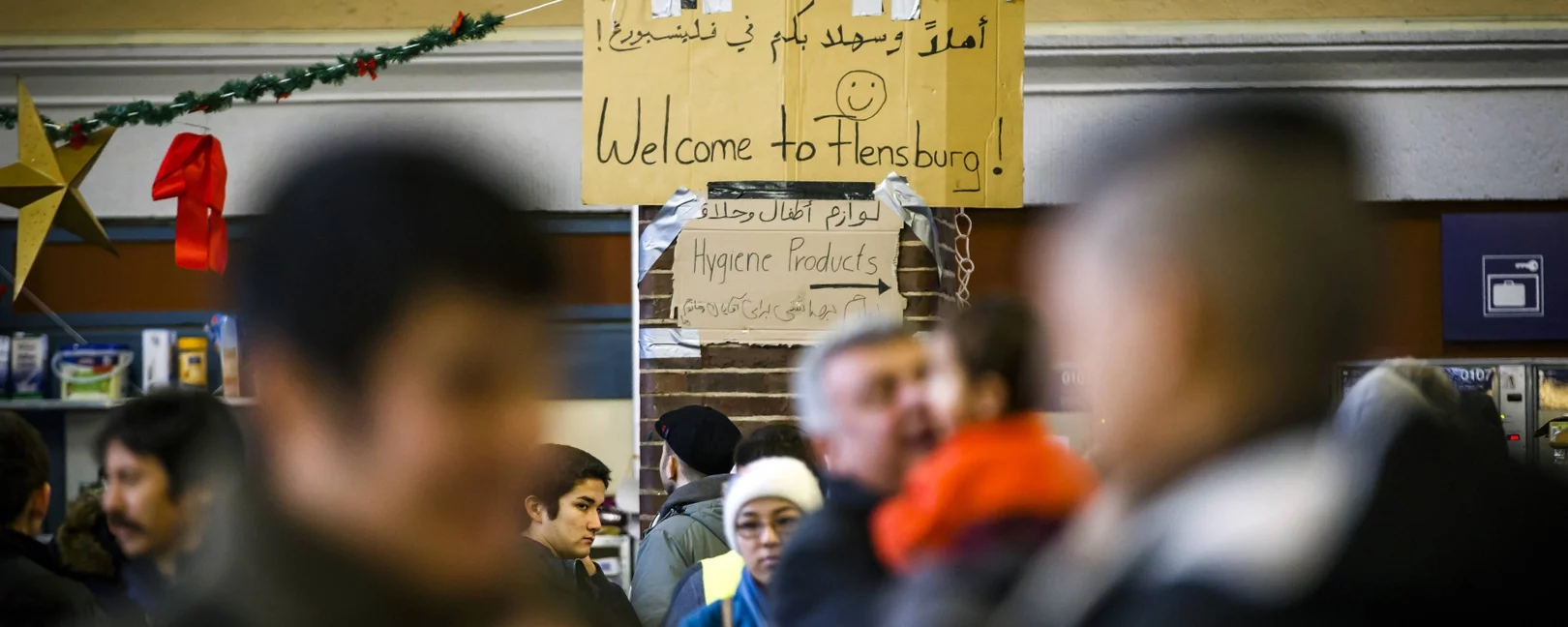 Mehrere Menschen in einer belebten Halle, im Hintergrund ein Schild mit „Welcome to Flensburg!“