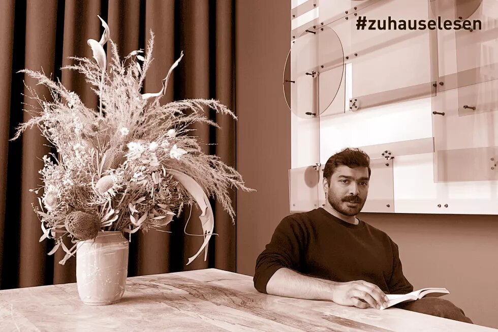 Senthuran Varatharajah sitzt an einem Tisch, auf dem ein großer Blumenstrauß steht, während seiner Video-Lesung