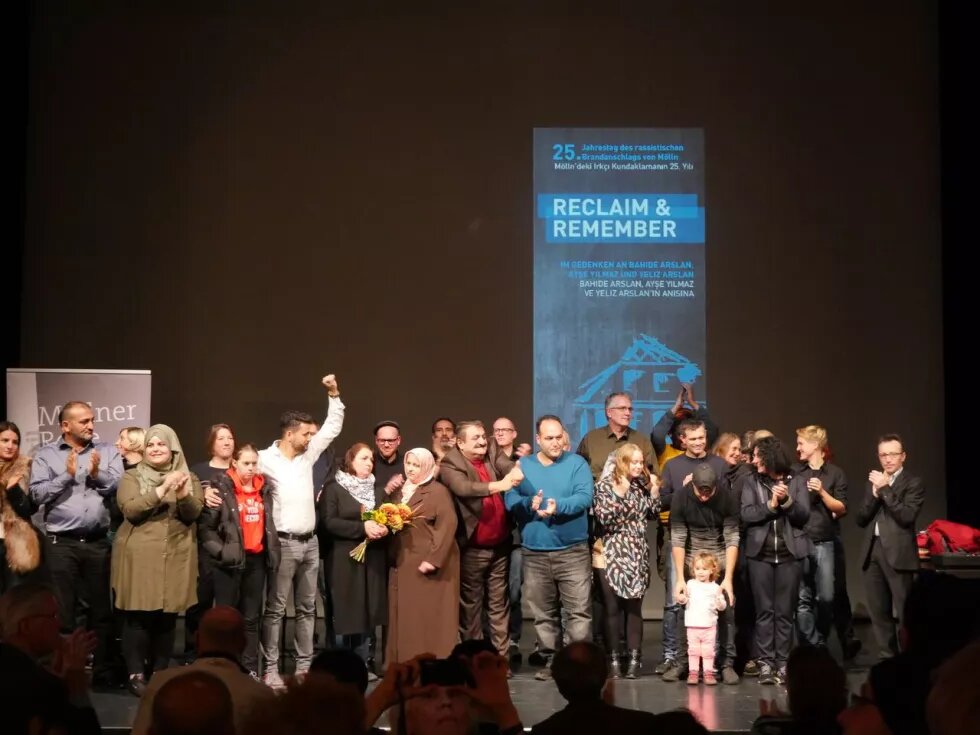 Möllner Rede im Exil - Menschengruppe steht auf einer Bühne, im Hintergrund ein Banner mit der Aufschrift "Reclaim and Remember"