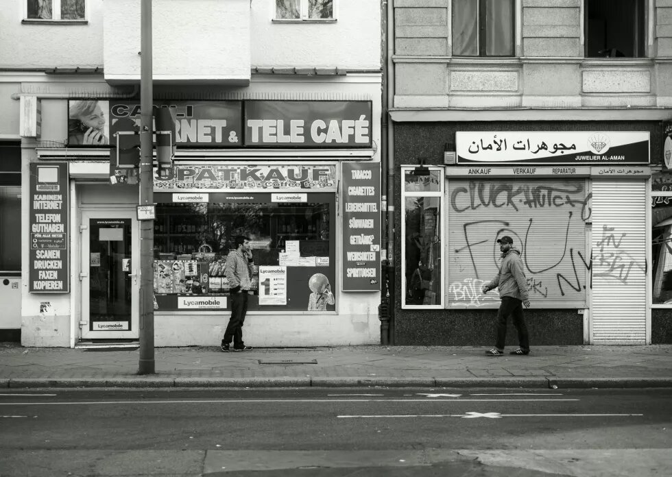 Schwarz-weiß-Fotografie einer Straße in Neukölln, zu sehen ist ein Spätkauf mit dem Schriftzug "Internet Tele Cafe", vor dem ein rauchend ein Mann steht, daneben ein Juweliergeschäft mit arabischem Schriftzug