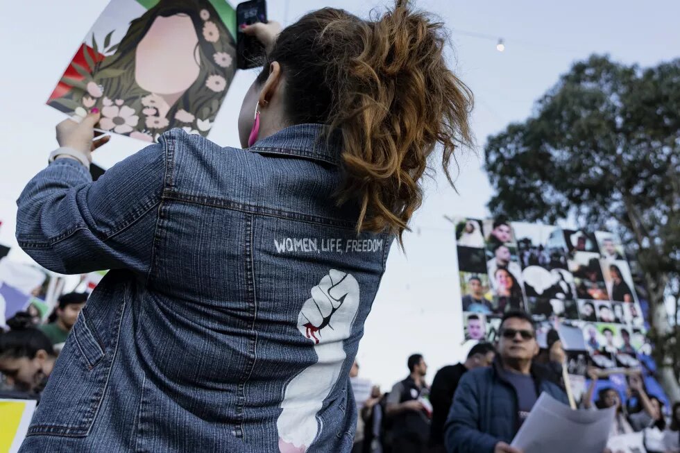 Eine Frau auf einer Solidaritätskundgebung in Melbourne hält ein Bild mit der Silhouette einer Frau mit langen, offenen Haaren hoch, im Hintergrund sind die Farben der iranischen Flagge zu sehen. Auf dem Rücken ihrer Jacke ist eine erhobene Faust zu sehen, darüber steht "Women, life, Freedom"