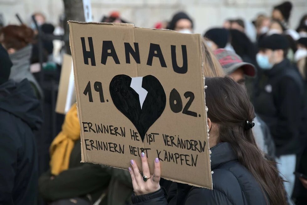 Bei einer Gedenkveranstaltung am 19.02.21 wird ein Schild hochgehalten mit einem schwarzen Herz in der Mitte, durch das oben ein weißer Riss geht. Darüber steht Hanau 19.02., darunter Erinnern heißt Verändern - Erinnern heißt Kämpfen