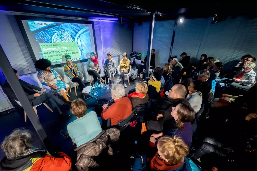 Vier Personen sitzen auf einer Bühne bei einer Podiumsdiskussion zum Thema  "Ostdeutsch-Plus: Die DDR-Migrationsgesellschaft seit dem Fall der Mauer", im Vordergrund ist Publikum zu sehen