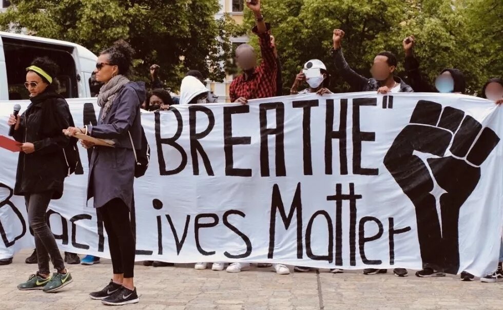 Bild von einer BLM Demo in Magdeburg, zwei Personen stehen vor einem Banner, auf dem Black Lives Matter steht und eine erhobene Faust zu sehen ist