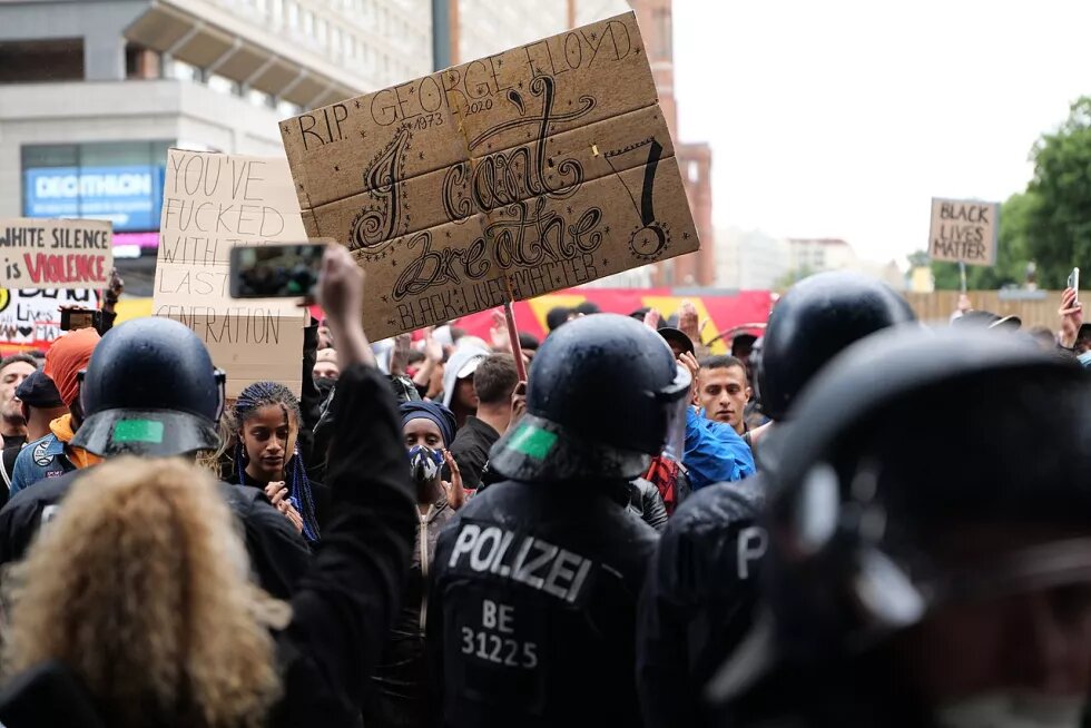 Black Lives Matter Demonstration in Berlin im Juni 2020: zu sehen sind behelmte Polizist*innen im Vordergrund, im Hintergrund Demonstrierende, eine Person hält ein Schild hoch mit der Aufschrift "RIP George Floyd, I can't breathe, Black Lives Matter"
