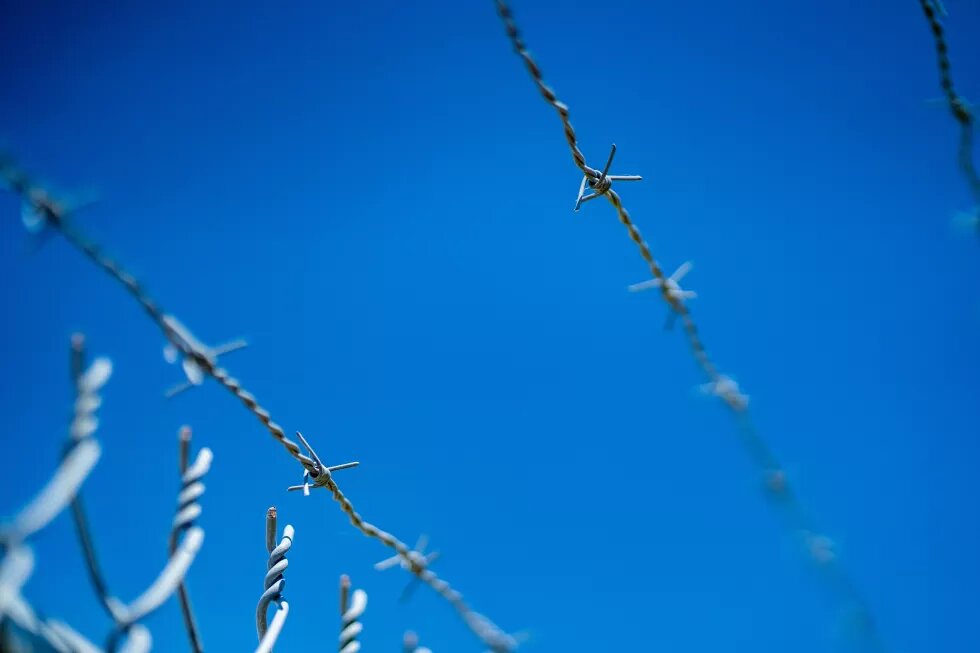 Vor strahlend blauem Himmel ist ein Stacheldrahtzaun zu sehen, schräg von unten fotografiert.