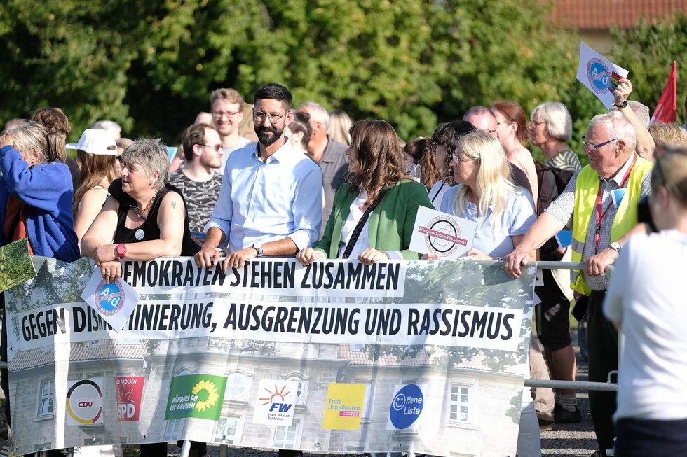 Mehrere Menschen halten bei einer Kundgebung einen großen Banner in den Händen, auf dem "Demokraten stehen zusammen gegen Diskriminierung, Ausgrenzung und Rassismus" steht; darunter sind die Logos folgender Parteien platziert: CDU, SPD, FW, FDP, Offene Liste. 