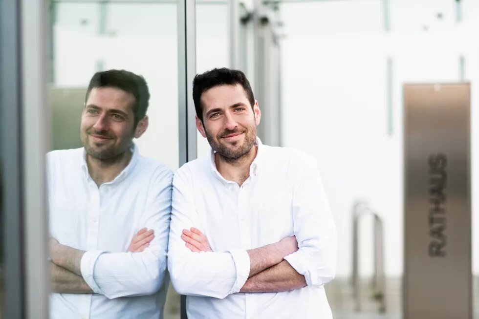 Portraitfoto von Ryyan Alshebl, der lächelnd an einer Glaswand gelehnt steht, in der er sich spiegelt. Im Hintergrund steht "Rathaus".