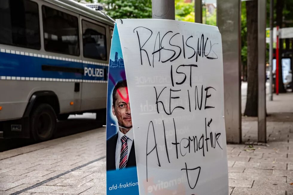 Foto eines umgedrehten Wahlplakats der AfD, auf dessen Rückseite "Rassismus ist keine Alternative!" geschrieben wurde, im Hintergrund ein Polizeiwagen.