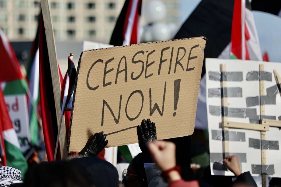 Bei einem Protest wird ein Schild mit der Aufschrift "Ceasefire Now!" hochgehalten, im Hintergrund sind palästinensische Flaggen zu sehen