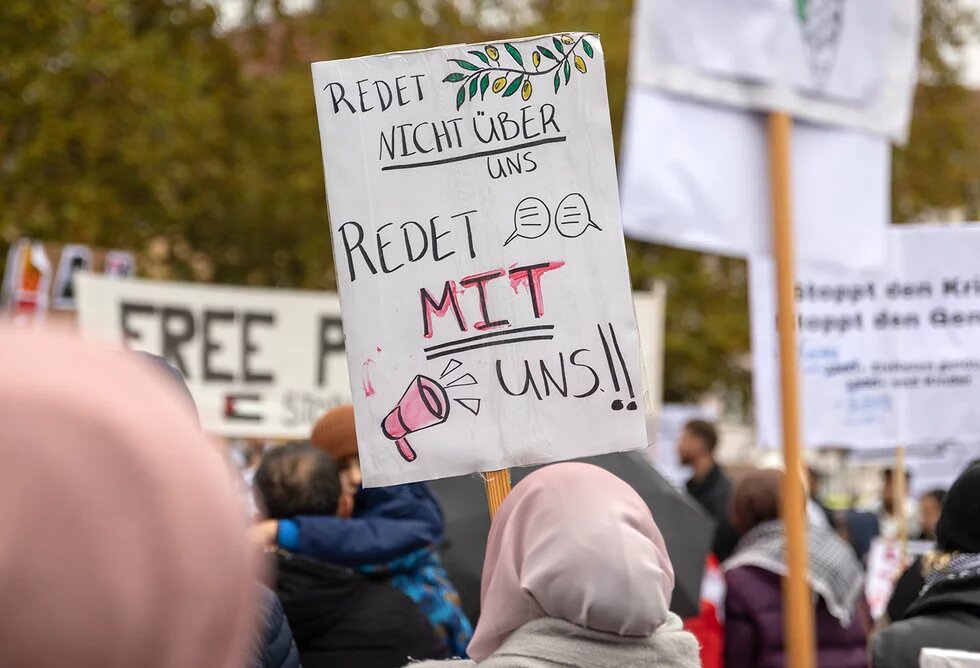 Bei einer Demonstration hält eine Person mit Kopftuch ein Schild hoch mit der Aufschrift "Redet nicht über uns, redet mit uns!!"