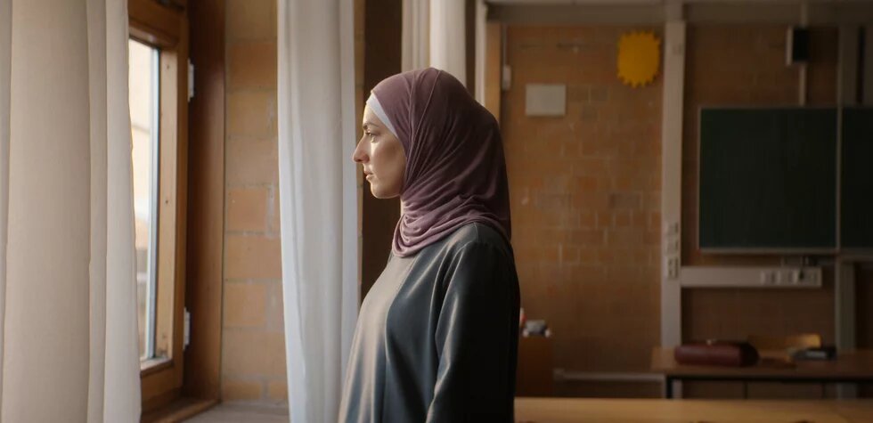 Filmstill aus der Episode "Nine Eleven" der Serie "Uncivilized": Eine junge Frau mit Kopftuch steht in einem Klassenzimmer und schaut aus dem Fenster