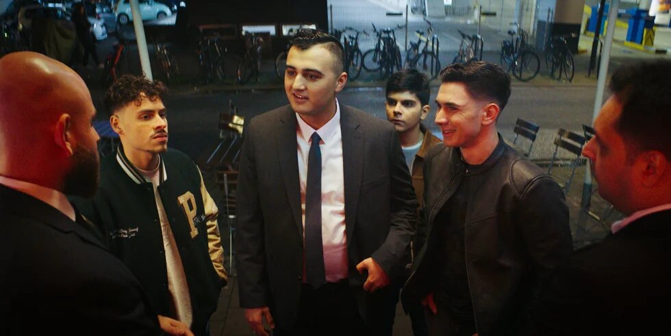 Filmstill aus der Episode "Hanau" von Uncivilized: Vier migrantische männliche Jugendliche, einer davon mit Anzug und Krawatte, stehen vor zwei Türstehern.