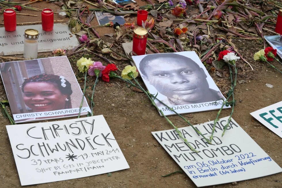 Zwischen Grabkerzen und Blumen liegen Bilder von zwei Menschen, darunter steht jeweils "Christy Schwundeck" und " Kupa Ilunga Medard Mutombo" 