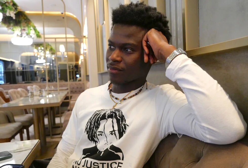 Suraj Mailitafi sitzt in einem Café, er trägt ein weißes T-Shirt mit einem Bild von Lorenz A.