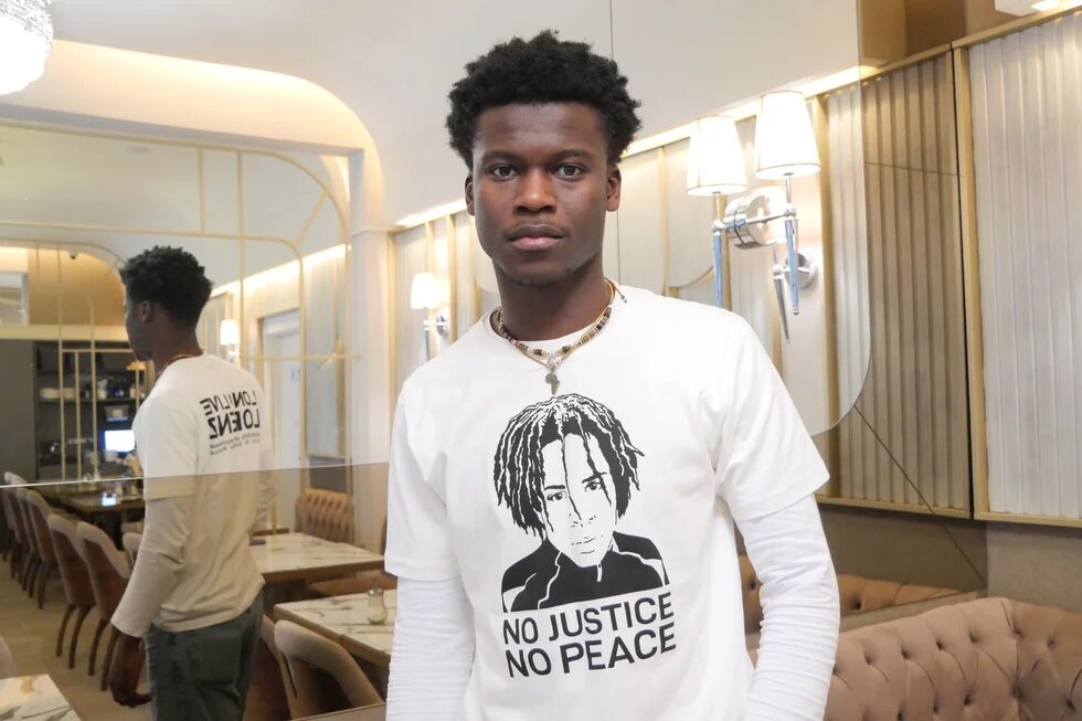 Suraj Mailitafi schaut in die Kamera und trägt ein weißes T-Shirt mit einem Bild von Lorenz und den Worten "No Justice, No Peace". 