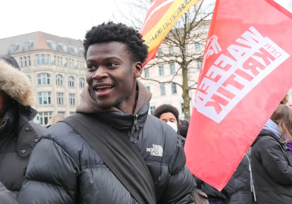 Suraj Mailitafi läuft in einer Menschengruppe auf einer Demo, neben ihm wehen zwei rote Fahnen