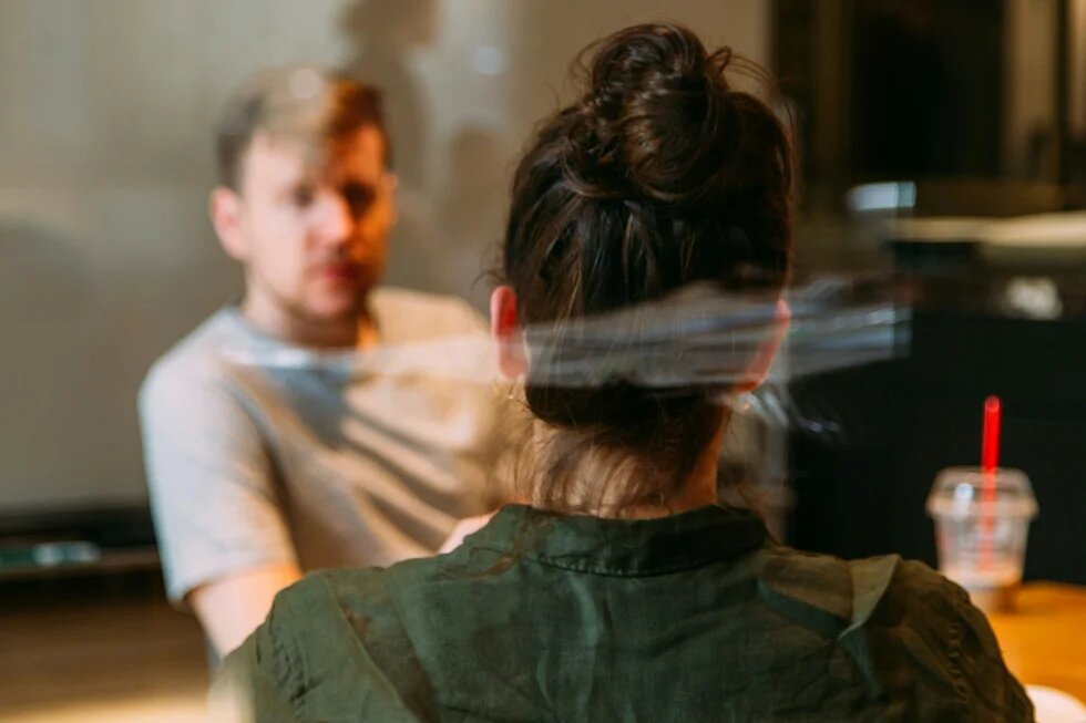 Zwei Personen sitzen an einem Tisch: Eine Frau, die von hinten zu sehen ist, und ein Mann, der vor ihr sitzt und sie anschaut