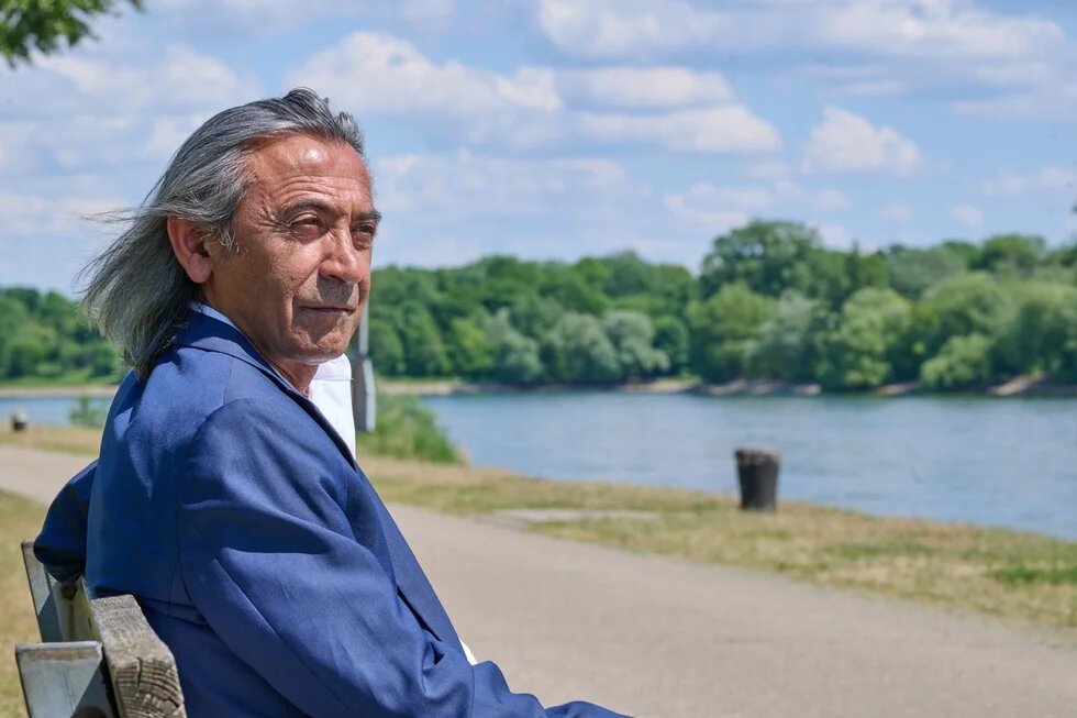 Ein Mann sitzt auf einer Bank an einem Fluss und schaut in die Ferne.