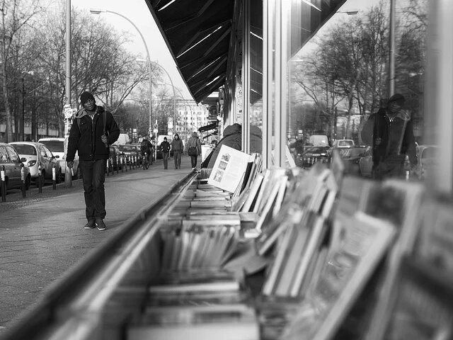 Strassenaufnahme im Vordergrund unscharf Kisten mit Büchern vor einem Geschäft, im Hintergrund ein Mann dessen Bild sich in der Scheibe des Geschäfts spiegelt