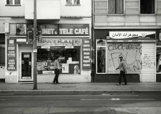 Berlin Neukölln Schwarz-weiß-Fotografie einer Straße in Neukölln, zu sehen ist ein Spätkauf mit dem Schriftzug "Internet Tele Cafe", vor dem ein rauchend ein Mann steht, daneben ein Juweliergeschäft mit arabischem Schriftzug