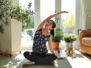 Lina Lam sitzt in einem lichtdurchfluteten Zimmer auf einer Yoga-Matte im Schneidersitz und macht eine Dehnübung mit ihrem rechten Arm