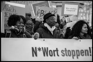 Bei einer Demo halten Schwarze Menschen einen Banner mit der Aufschrift "N*Wort stoppen!", im Hintergrund sind weitere Demoschilder zu sehen, mit der Aufschrift "#Black" oder "Wir erheben Einspruch".