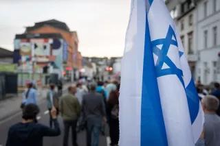Bei einem Schweigemarsch in Solidarität mit Israel in Saarbrücken sind auf einer Straße laufende Menschen von hinten zu sehen, im Vordergrund eine Israel-Flagge.