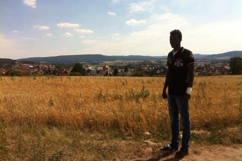 junge Geflüchtete in Kleinstadt in Bayern Junger Mann auf einem Feld, unter blauem Himmel, vor einer Kleinstadt und Wäldern